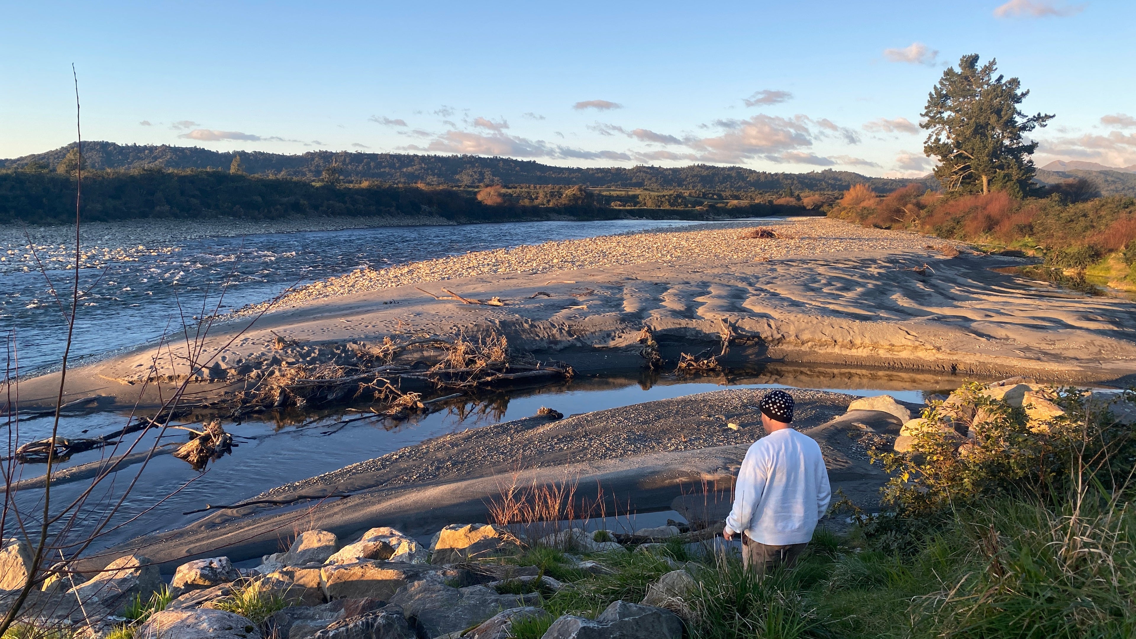 Person standing on a rocky shoreline with a scenic view of water and mountains.